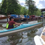 Kajakfahrer paddelt durch einen ruhigen Kanal neben vertäuten Booten und Bäumen