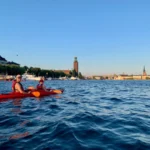 Sonrientes kayakistas remando frente al Ayuntamiento de Estocolmo durante una dorada tarde de verano. Una forma divertida y única de ver la ciudad desde el agua.