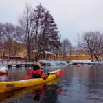 Piragüista solitario navegando por un tranquilo canal rodeado de árboles desnudos y orillas nevadas.