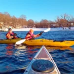 Dos kayakistas ataviados con cálidos trajes secos remando por las gélidas aguas de Estocolmo con árboles nevados al fondo.